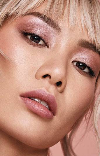 closeup of model's face. She has blonde hair, short bangs and is wearing rose gold-hued makeup.